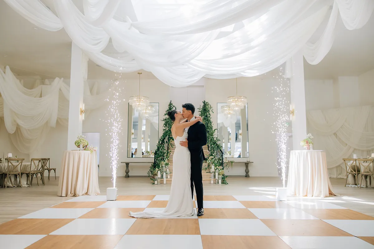 Couple kissing on the checkered dance floor under the ceiling draping, spark fountains and chandeliers behind them
