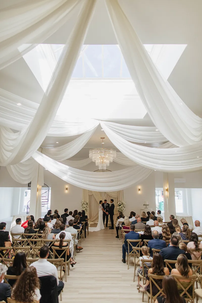 Guests seated for a ceremony beneath ceiling draping and a chandelier, the couple at the altar under the skylight