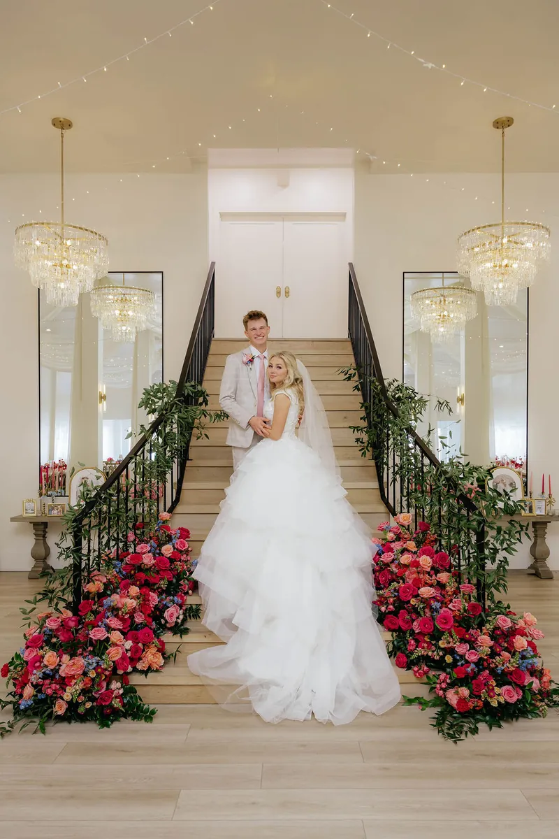 Bride and groom on the staircase framed by pink and coral florals under gold chandeliers